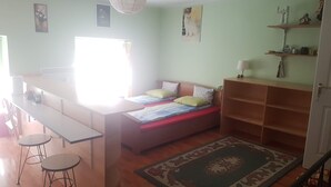 A room with a bed, a desk, two stools, a lamp, a shelf with items, and a window.