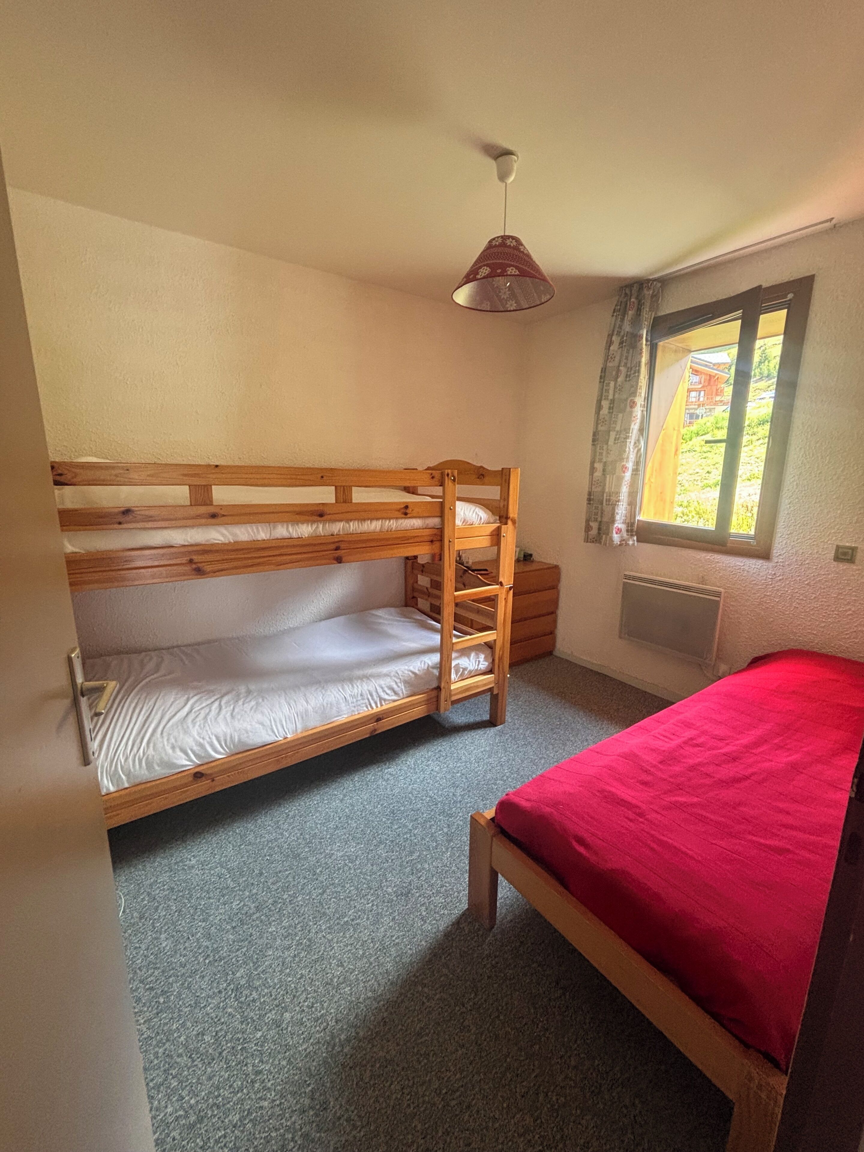 A bunk bed room with a red bedspread, a window with curtains, and a ceiling fan.