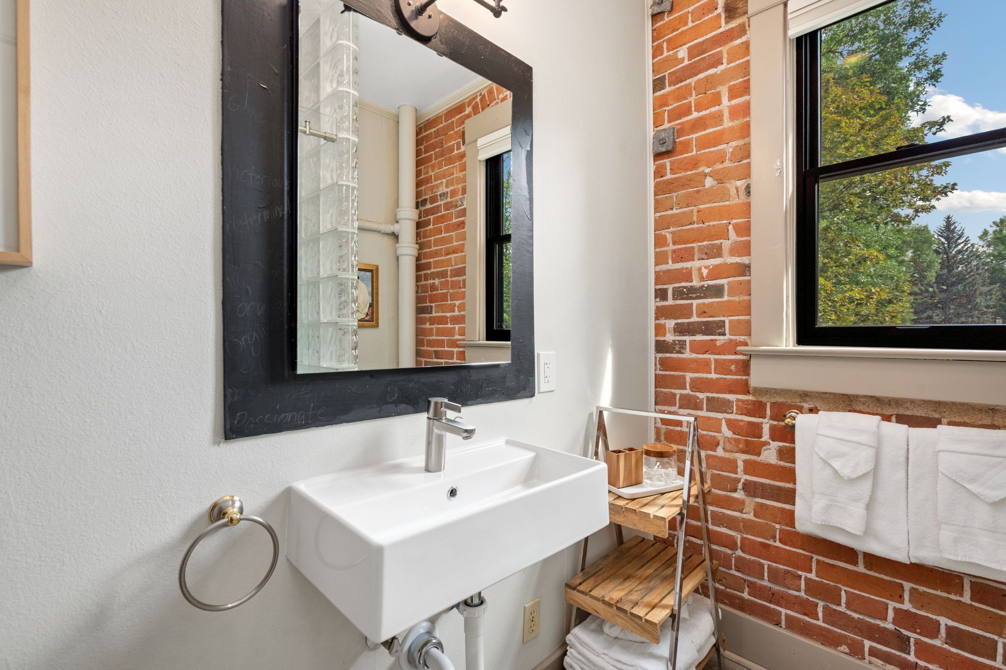 Un baño con lavamanos blanco, espejo y ventana con vistas a árboles.