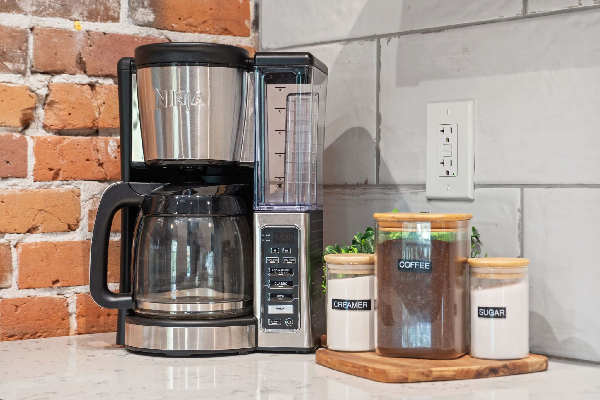 A coffee maker with a digital display and a timer, next to a brick wall and a power outlet.