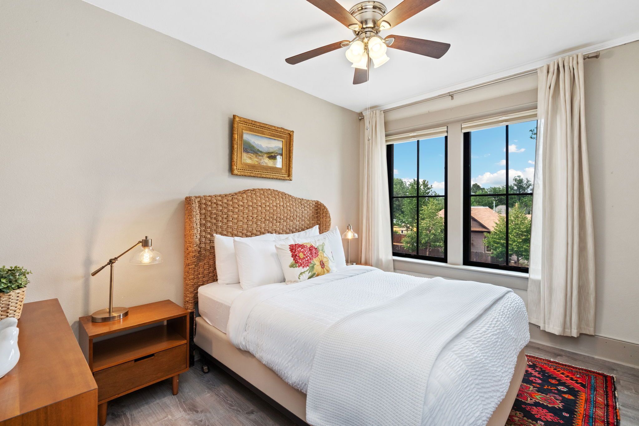 A bedroom with a bed, nightstands, a ceiling fan, a window with curtains, and a view of the outside.