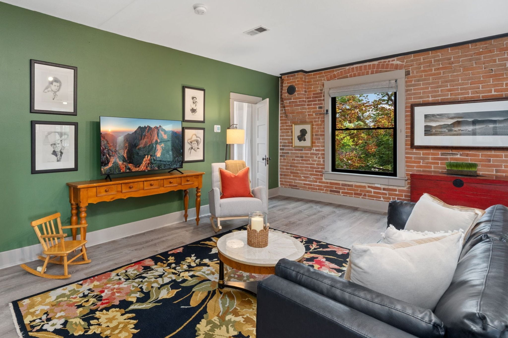 Una sala de estar con pared verde, pared de acento de ladrillo, un sofá, una mesa de centro, un televisor, una chimenea y una ventana con vistas.