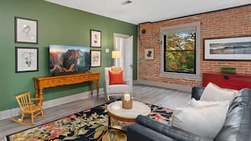 A living room with a green wall, a brick accent wall, a sofa, a coffee table, a TV, a fireplace, and a window with a view.