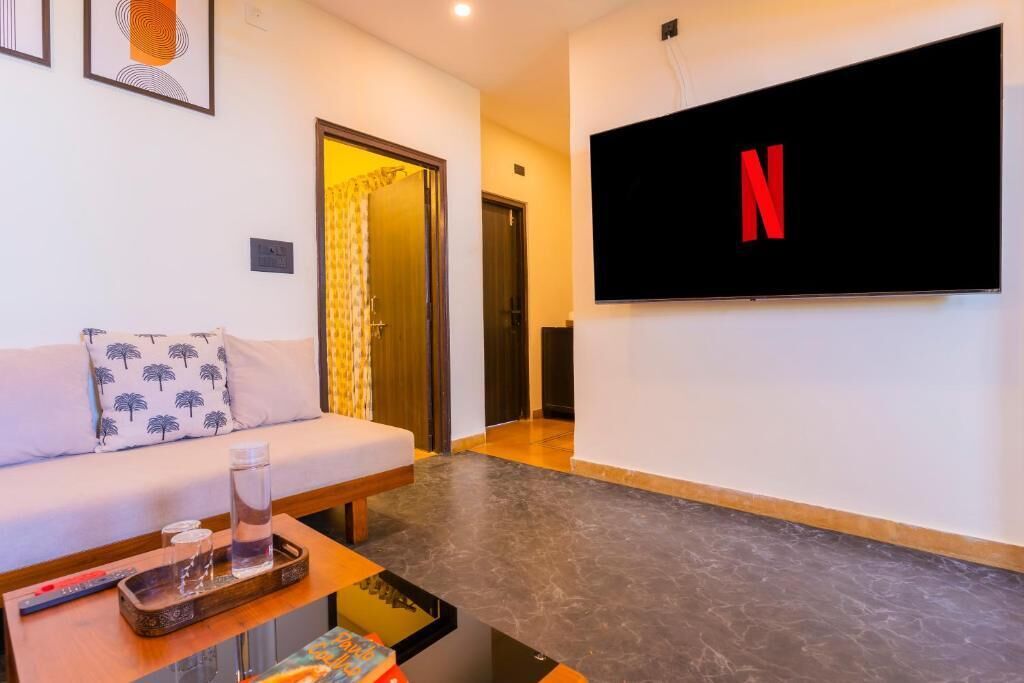 A living room with a sofa, a coffee table, a flat-screen TV mounted on the wall, and a door leading to another room.