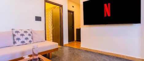 A living room with a sofa, a coffee table, a flat-screen TV mounted on the wall, and a door leading to another room.
