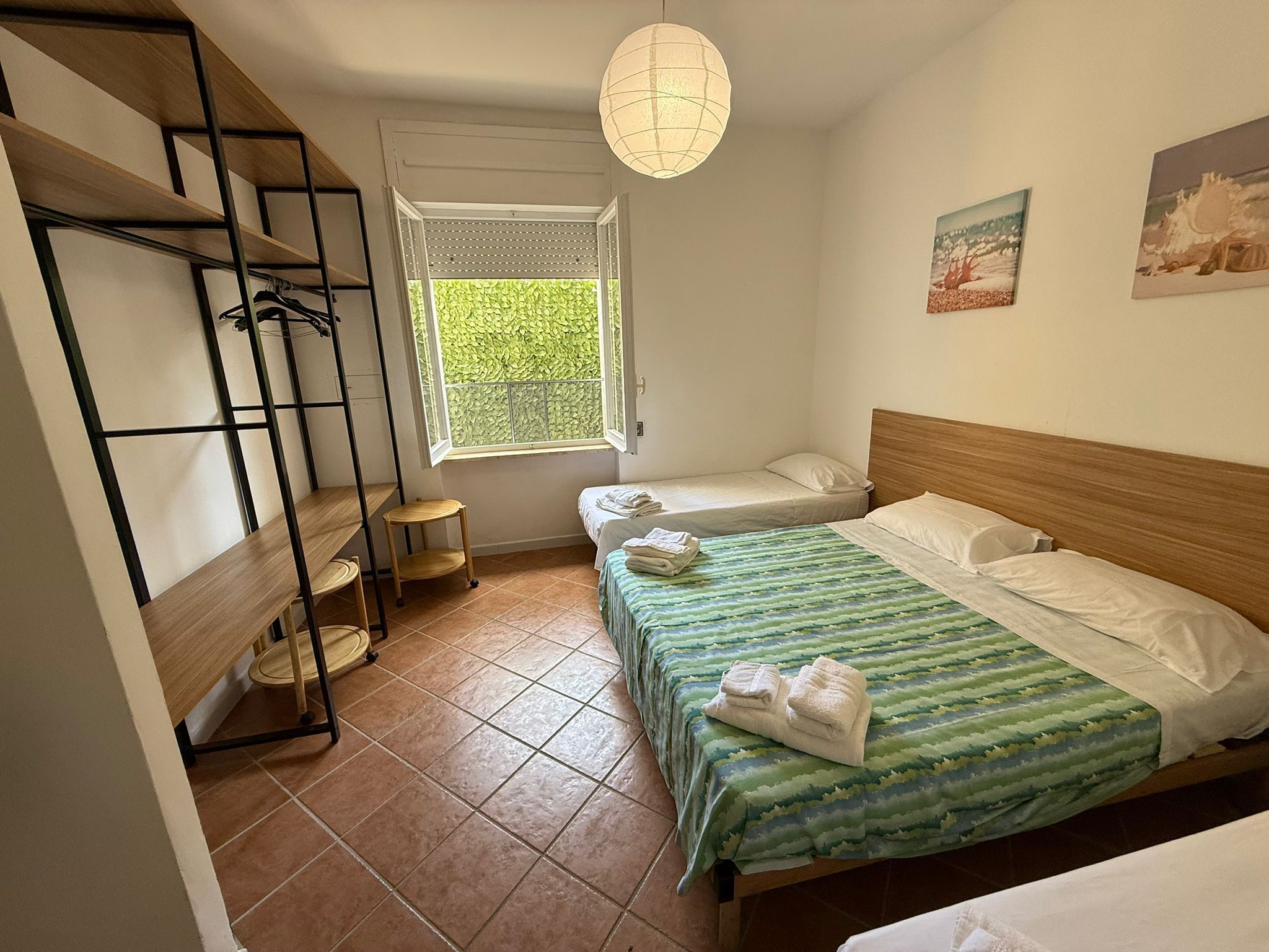 A bedroom with a bunk bed, a single bed, a window with blinds, a hanging light fixture, and a small bedside table.