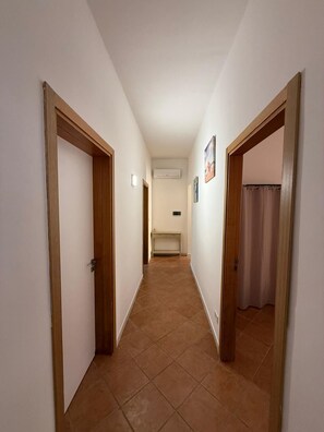 Um corredor estreito com portas de madeira em ambos os lados, piso ladrilhado e um aparelho de ar condicionado fixado na parede.