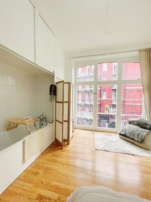 A room with a bathtub, wooden floor, a window with a view of red buildings, and a sofa with cushions. - Stylish Retreat at Hyde Park Mansions (London)