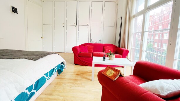 Ein Schlafzimmer mit einem Bett, einem roten Sofa, einem kleinen Tisch und einem Fenster mit Blick auf Gebäude.