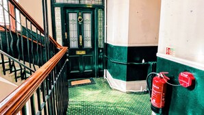 Eine Holzhandlauf-Treppe führt zu einer grünen Tür mit Glasfenster und Messinggriff. Ein roter Feuerlöscher ist an der rechten Wand angebracht.