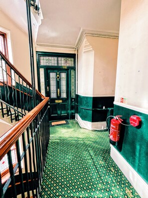 A staircase with a wooden handrail leading to a green door with a glass panel and a brass handle. A red fire extinguisher is mounted on the wall to the right. - Stylish Retreat at Hyde Park Mansions (London)