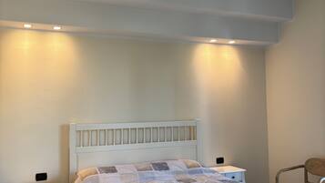 Una camera da letto con un letto bianco, un comodino, una sedia e una lampada a parete.