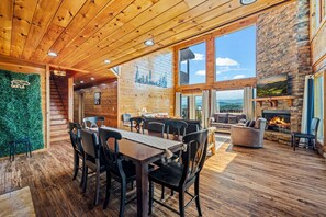 A spacious log cabin interior with a dining area, seating, a fireplace, and large windows offering scenic views. - Robin's View | Hot Tub, Indoor Pool, Unbeatable Mountain Views! (Pigeon Forge)