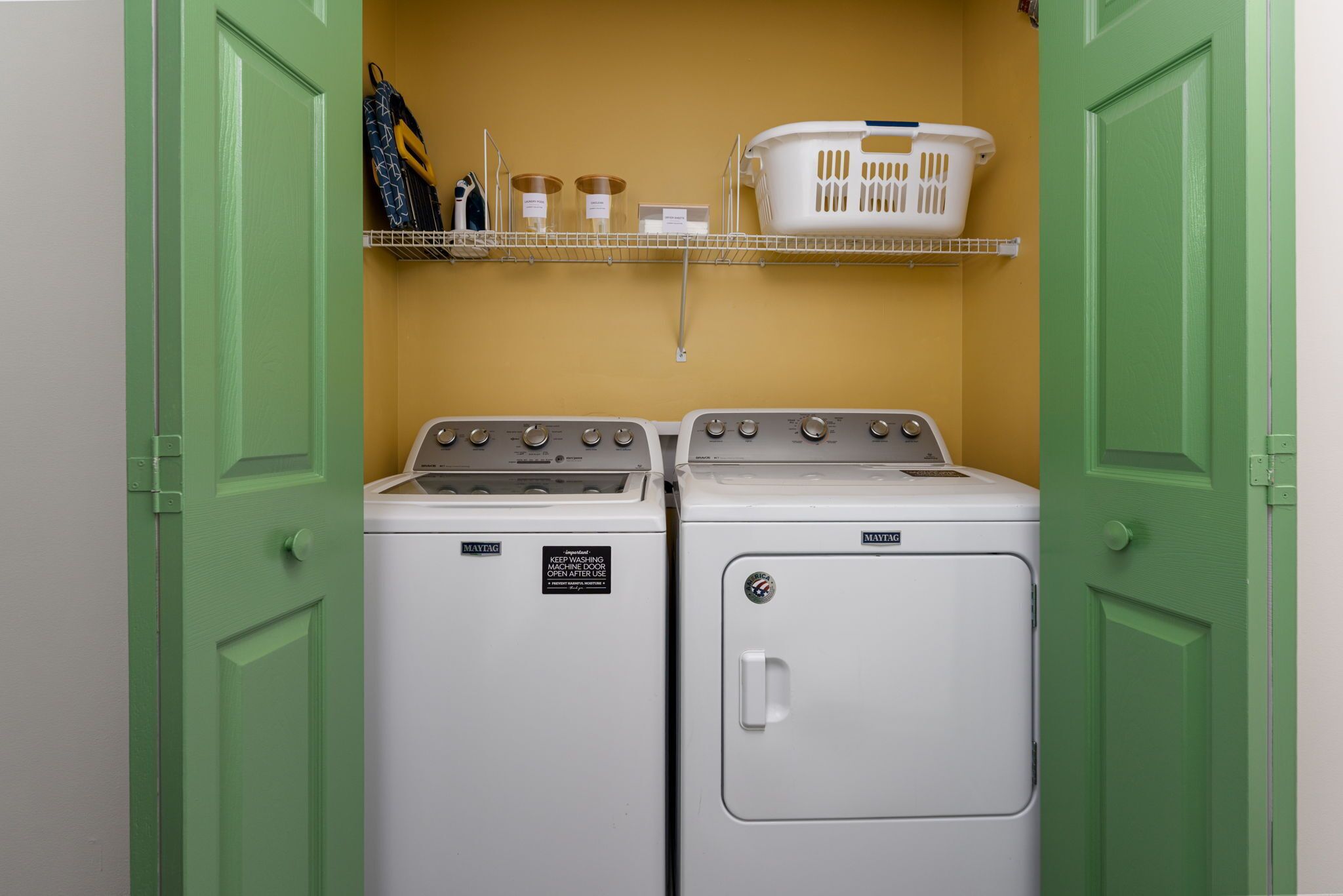 A laundry room with two Maytag washers and dryers, a shelf with laundry supplies, and a basket.