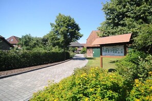 Un camí pavimentat que condueix a un edifici amb una placa verda, envoltat de vegetació i cases residencials.