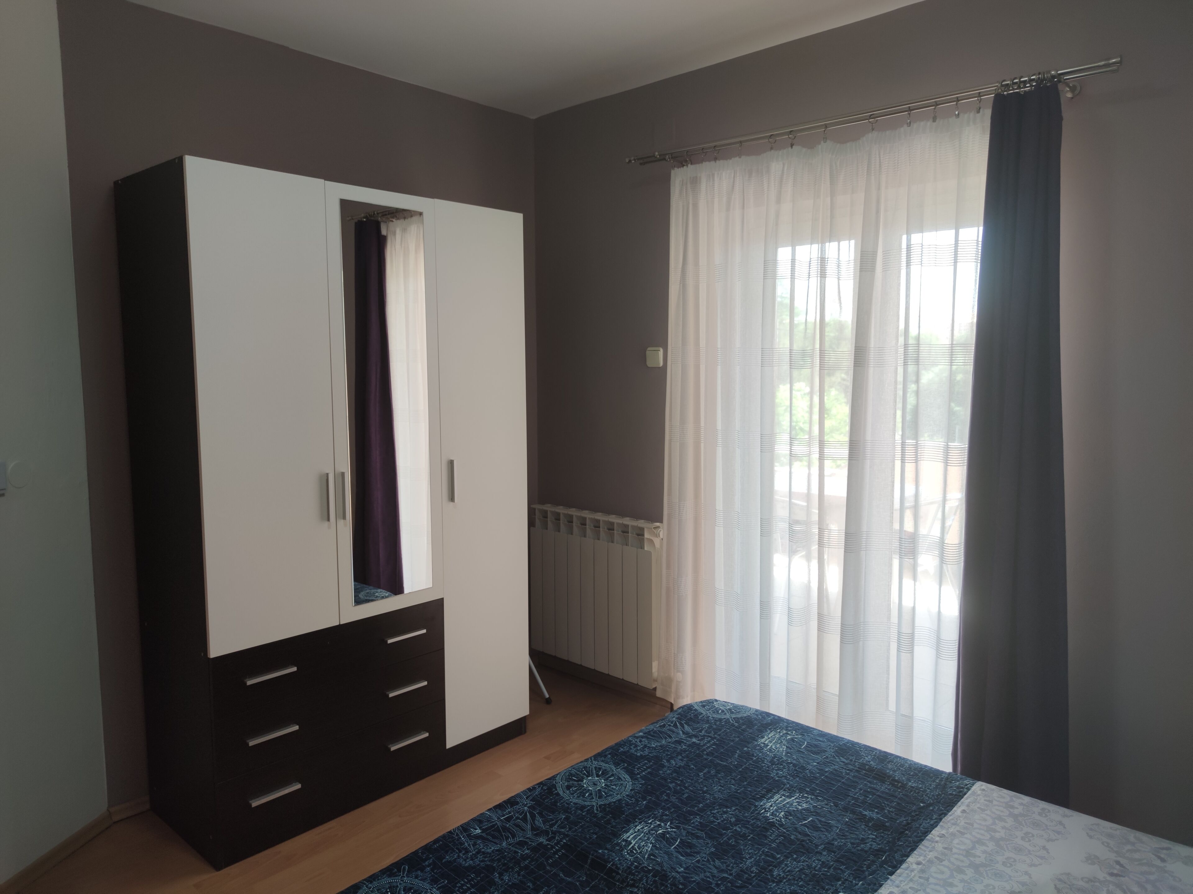A bedroom with a wardrobe, a bed with a patterned bedspread, and a window with curtains.