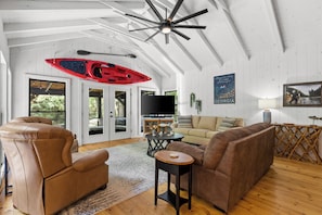 Ein geräumiges Wohnzimmer mit einer großen Couch, einem Ledersessel und einem Couchtisch. Es gibt einen Deckenventilator, einen wandmontierten Fernseher und ein rotes Kajak, das an der Wand hängt.