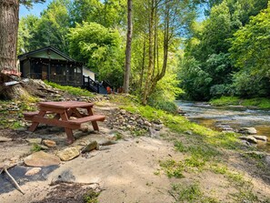 Ett rustikt picknickbord i trä vid en flod, omgivet av stenar och grönska, med en stuga i bakgrunden.