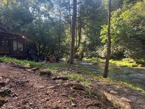Une cabane avec un espace extérieur aménagé, située près d’un ruisseau bordant une forêt.