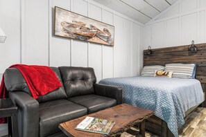 Een kamer met een leren bank, een houten salontafel met een tijdschrift, een bed met gestreept beddengoed en een schilderij aan de muur.