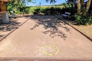 Uma mesa de jogo de shuffleboard com área de pontuação demarcada, rodeada por assentos externos e vegetação.
