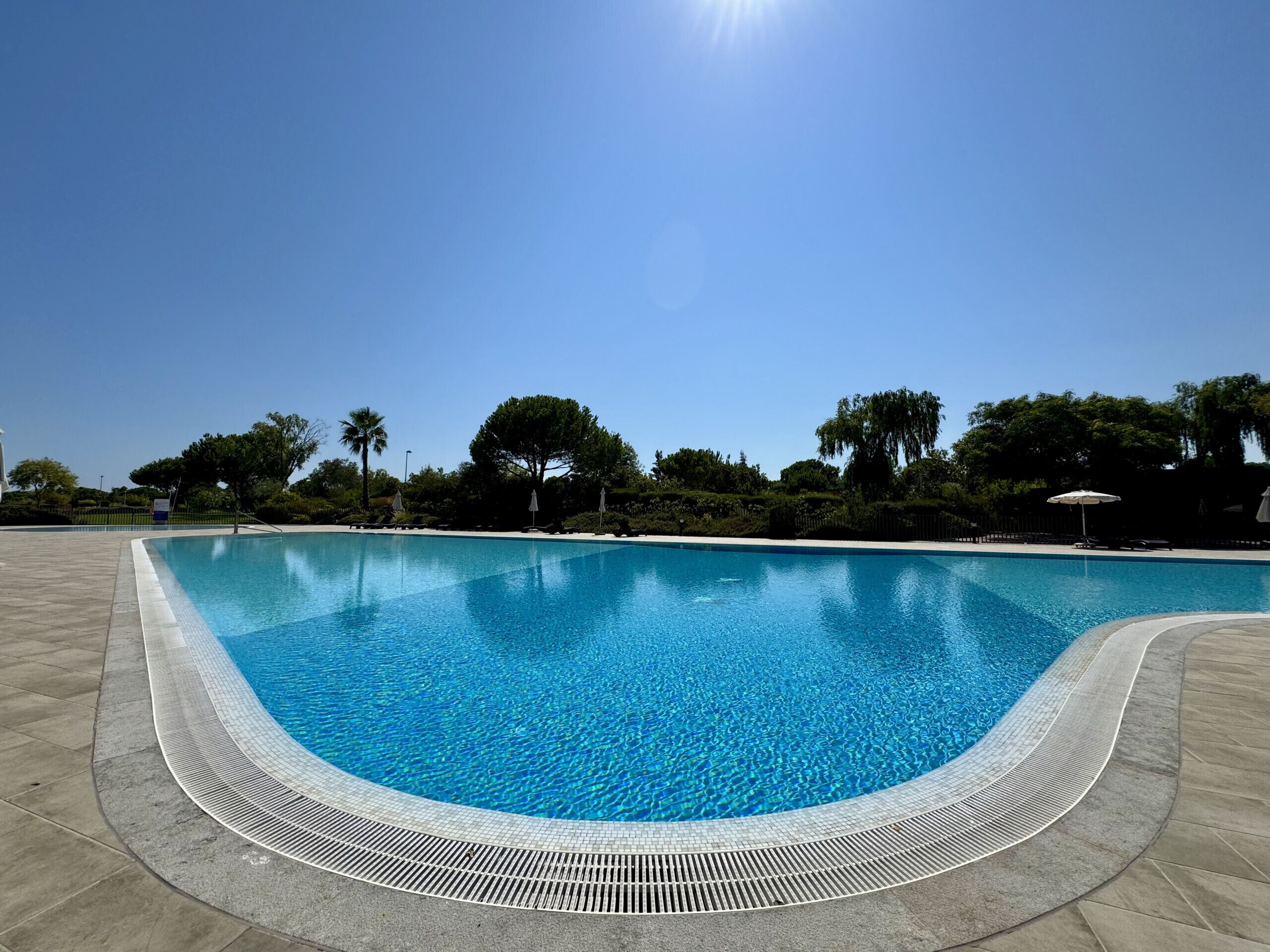 A clear blue pool with a tiled edge, surrounded by a paved area and trees.