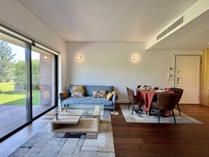 A modern living room with a glass sliding door leading to a patio, a blue sofa with patterned cushions, a glass coffee table, and a dining area with a red tablecloth. - Vilamoura Prestige 3 With Pool by Homing (Loulé)