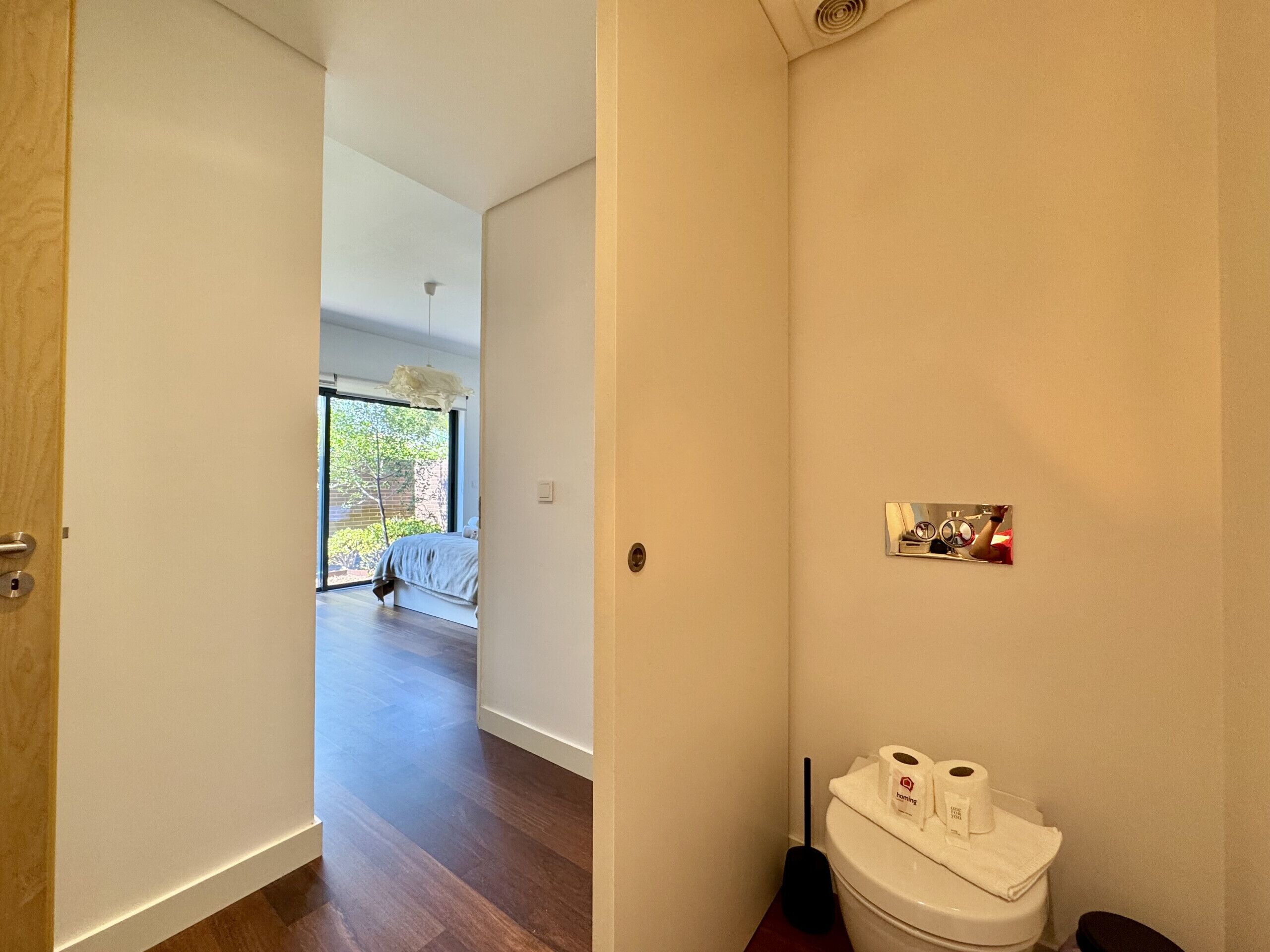 Un couloir menant à une chambre dotée d’une grande fenêtre, des toilettes avec du papier toilette et un balai.