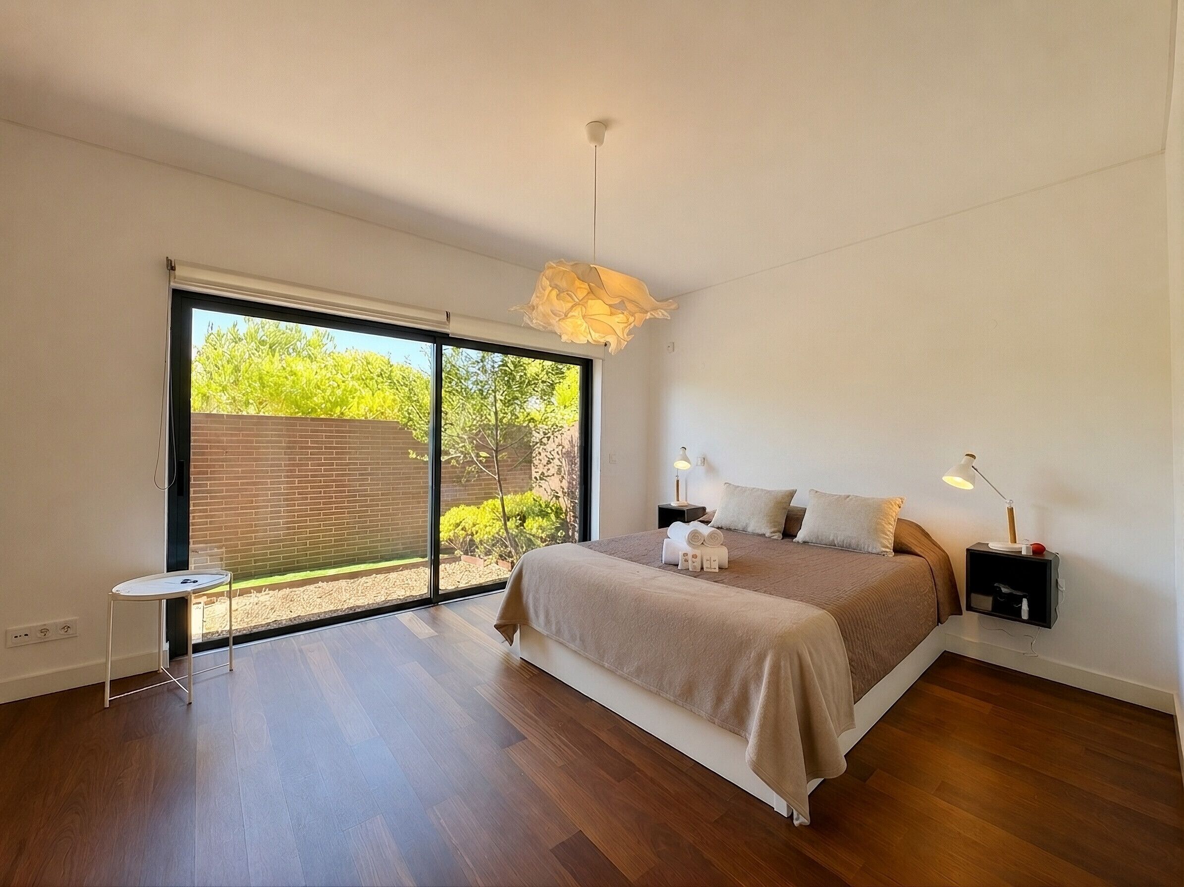 Un dormitorio con un amplio ventanal corredera que da a un jardín, una cama con cabecera, mesitas de noche y una lámpara colgante.