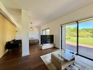 Sala de estar moderna com uma televisão grande, piso de madeira e vista para uma paisagem verde através de amplas janelas.