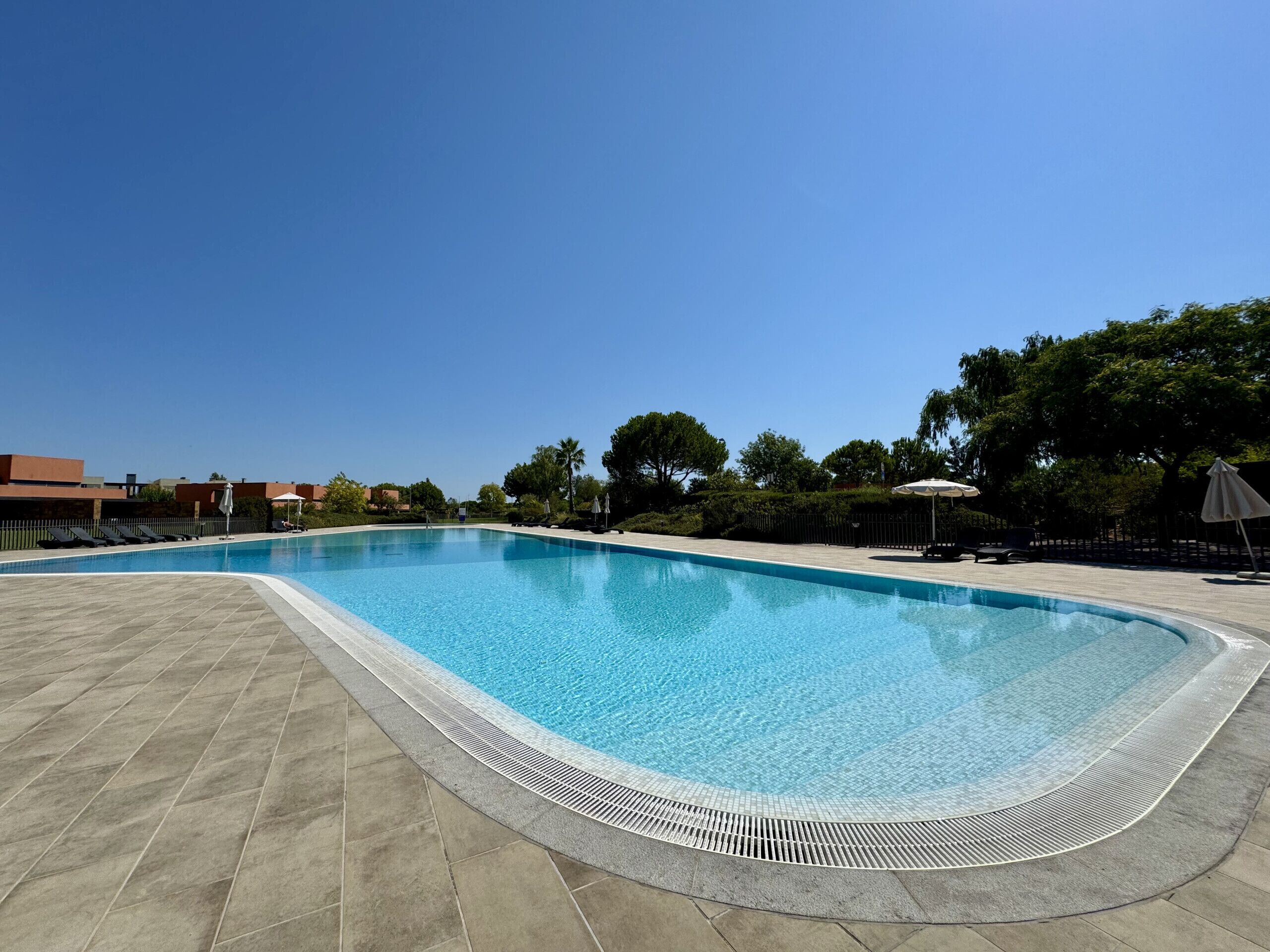 Una piscina rectangular con solárium alicatado, rodeada de sillones de playa y sombrillas, con árboles y edificios al fondo.