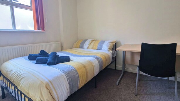 Uma cama de solteiro com um edredom listrado, uma mesa pequena, uma cadeira e uma janela com cortinas.