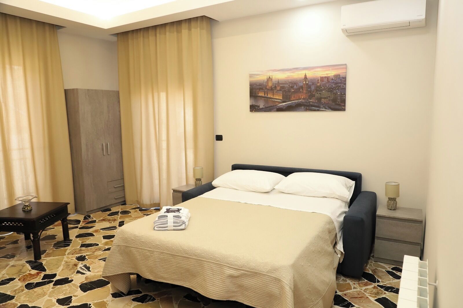 Une chambre d’hôtel comprenant un lit, une table de chevet, un bureau, une armoire et un tableau représentant un paysage urbain accroché au mur.