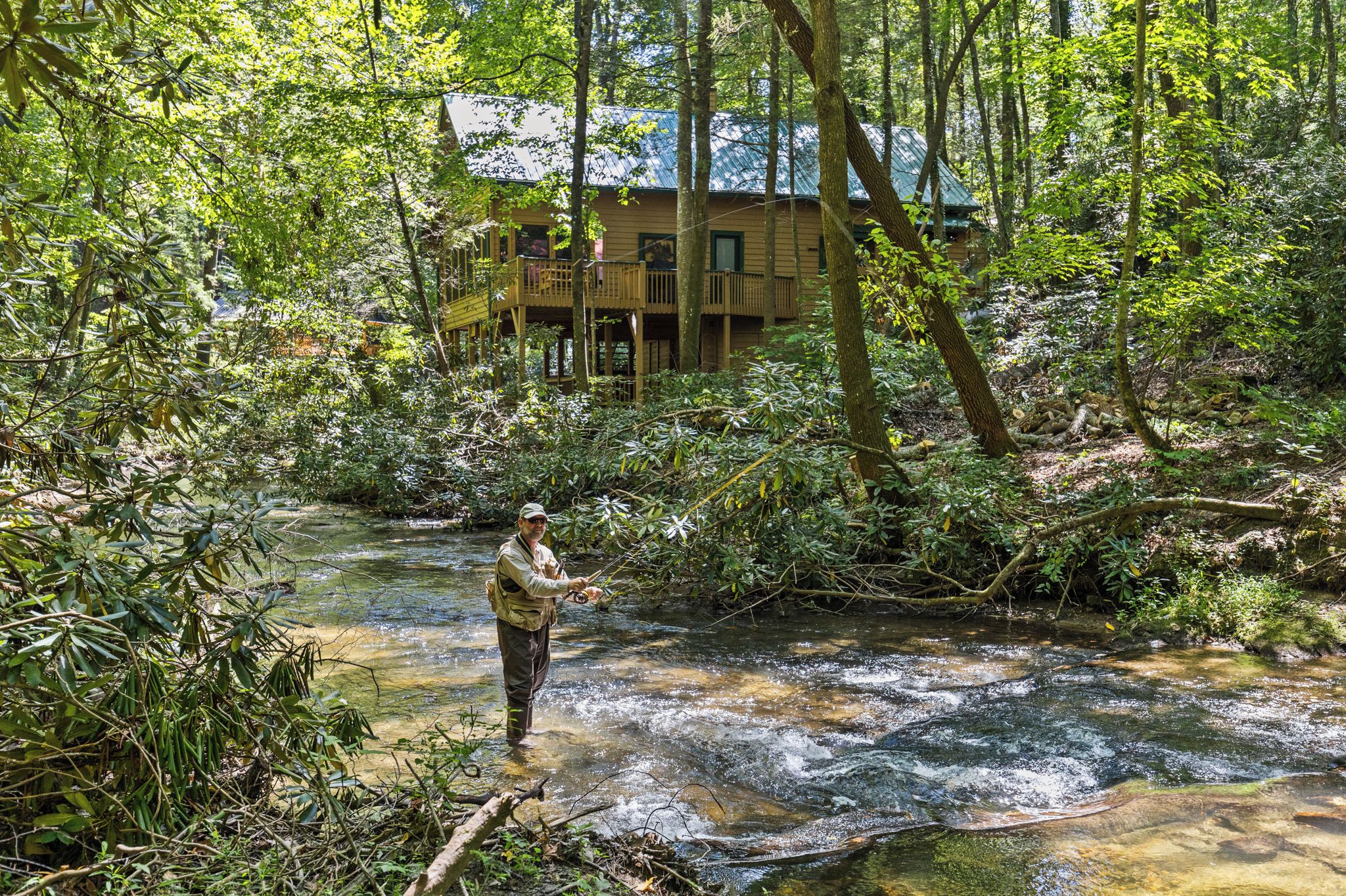 Un hombre está de pie en un arroyo en un bosque, con una casa al fondo.