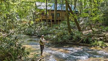 Un hombre está de pie en un arroyo en un bosque, con una casa al fondo.