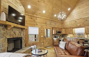 A wooden cabin interior with a stone fireplace, a television, a leather sofa, and a kitchen area.