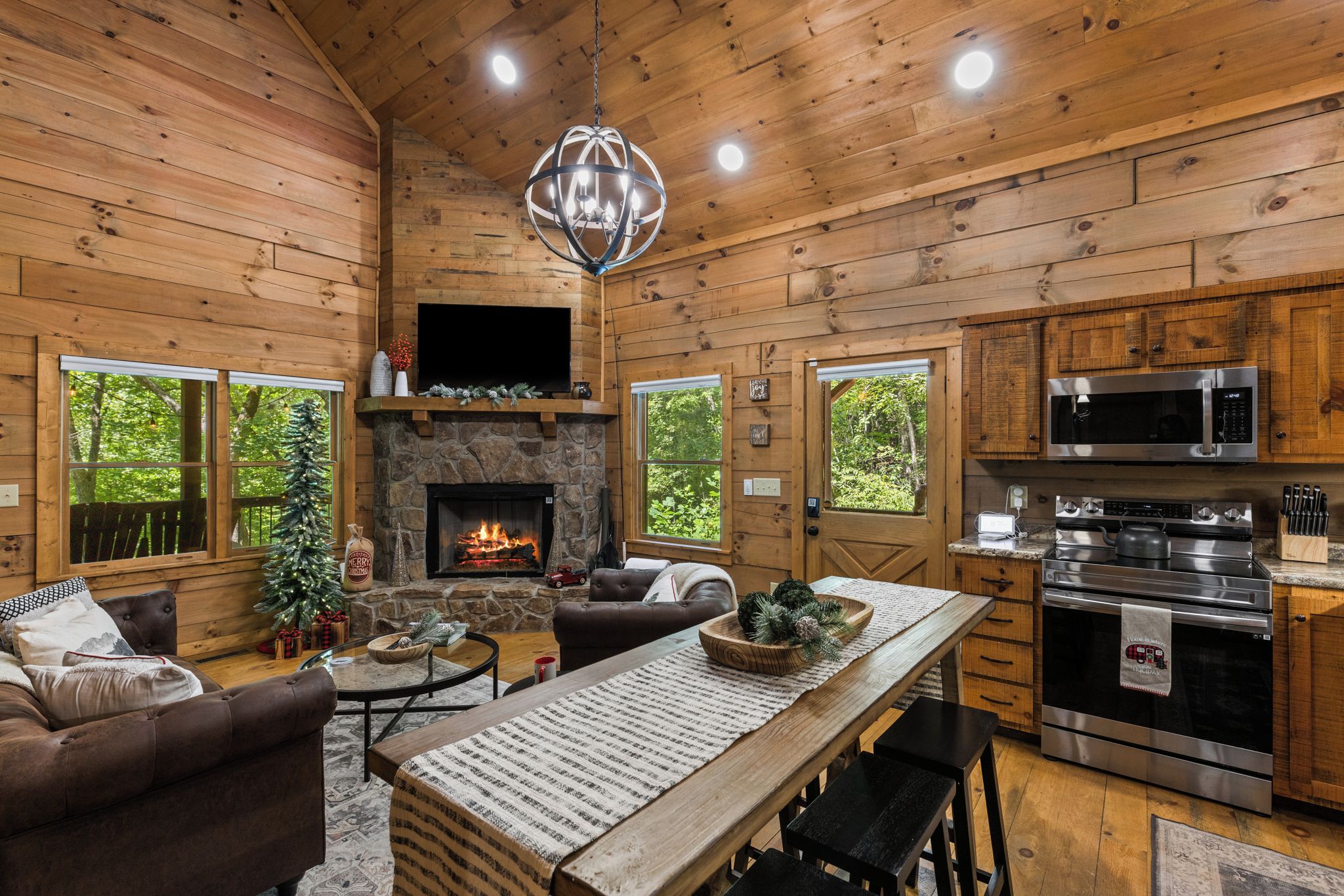 A cozy cabin interior with a stone fireplace, wooden walls, a dining area, and a kitchen with modern appliances.
