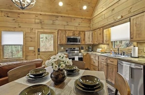 A wooden cabin interior with a dining table set for four, a kitchen with modern appliances, and a view of the forest through the windows.