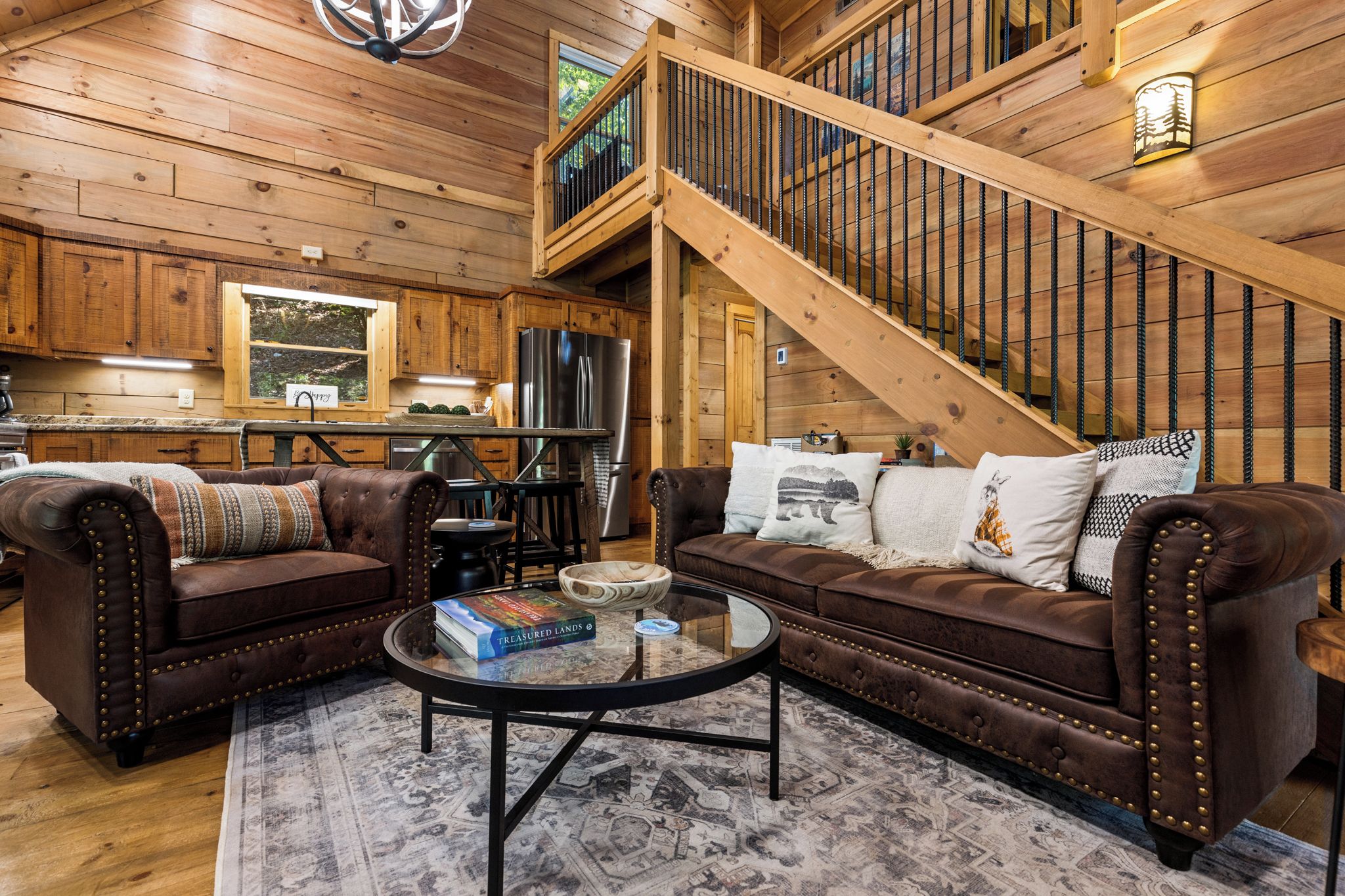 A cozy cabin interior with a wooden staircase, leather sofas, a dining area, and a kitchen.