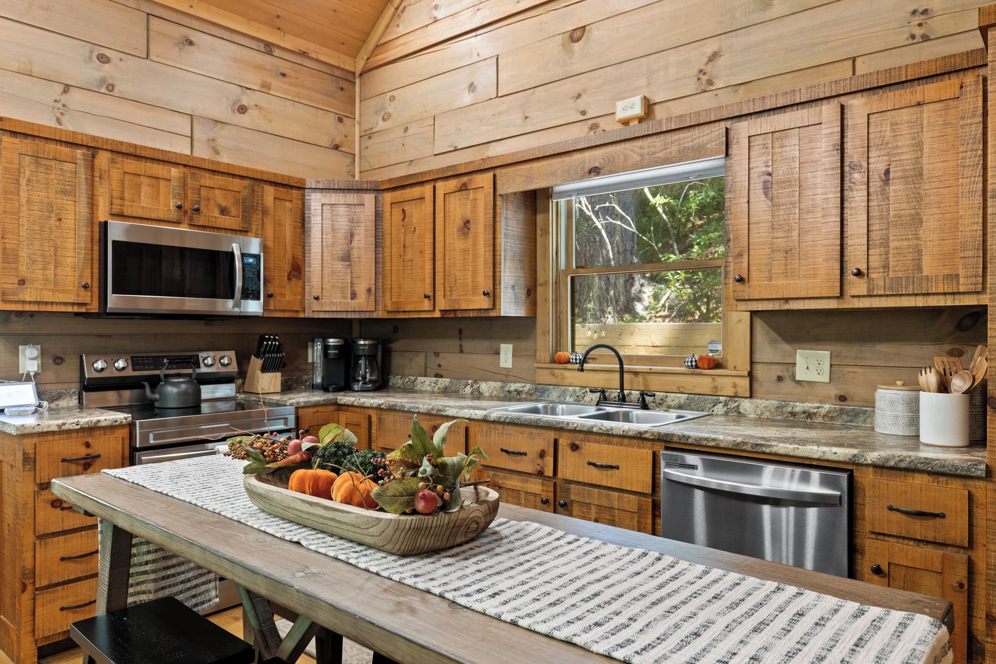 A kitchen with wooden cabinets, a microwave, stove, sink, and a window with a view of trees.