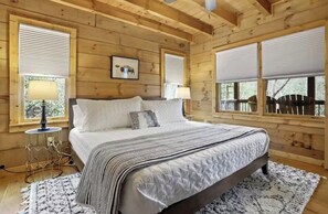 A wooden cabin bedroom with a bed, two bedside lamps, a window with blinds, and a picture on the wall.