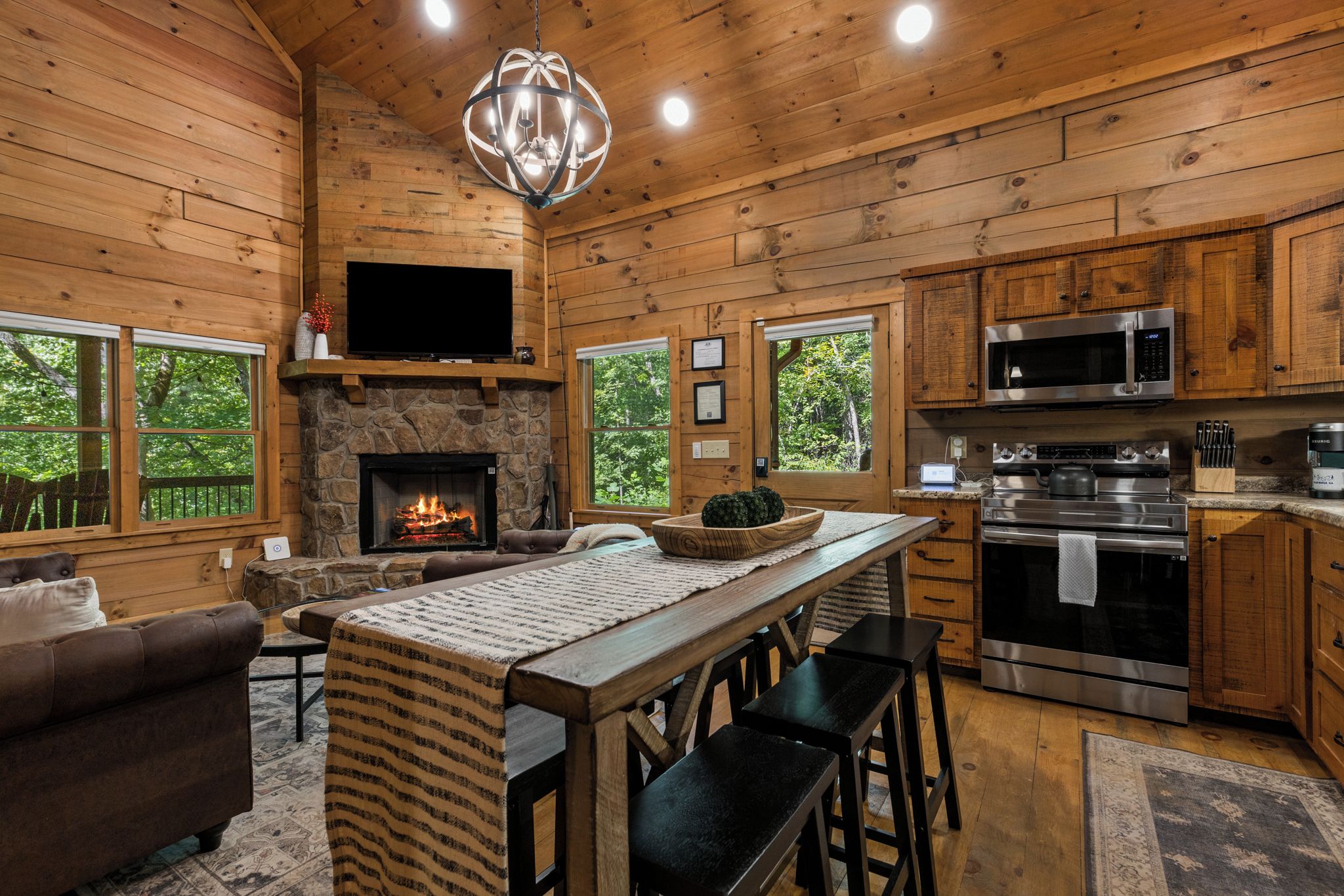 Un intérieur de chalet chaleureux avec une cheminée en pierre, des murs en bois, un téléviseur à écran plat et une cuisine moderne équipée d’appareils électroménagers en acier inoxydable.