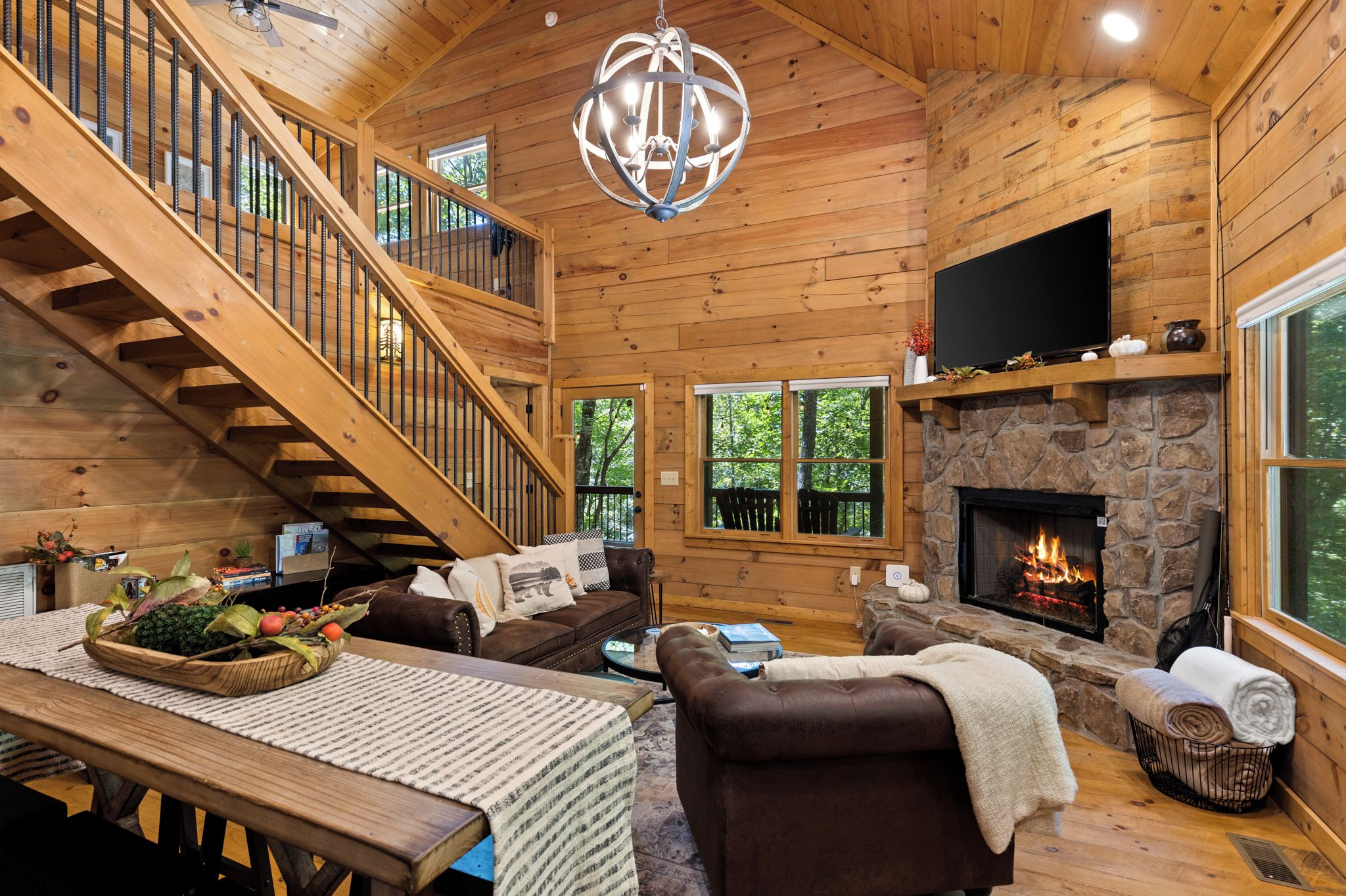 A log cabin interior with a stone fireplace, wooden staircase, and a television mounted above it.