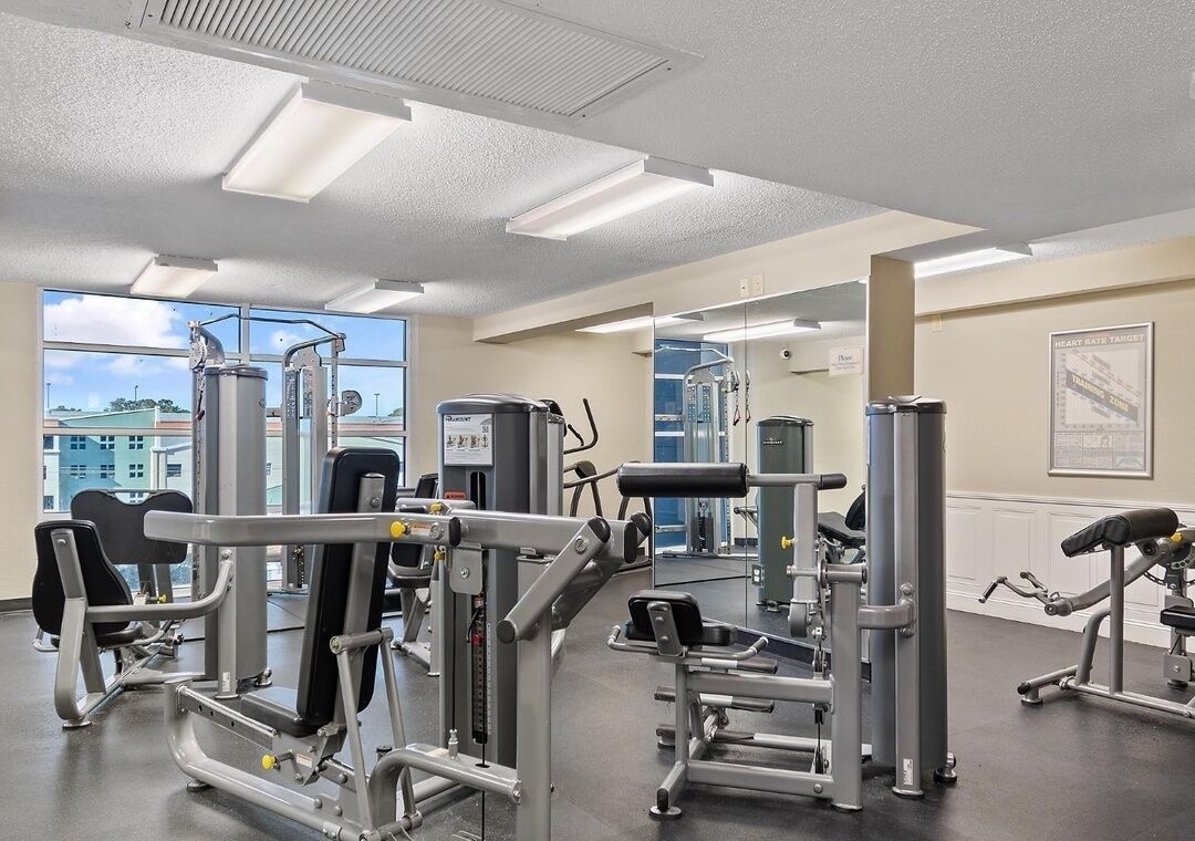 Un gimnasio con diferentes máquinas de ejercicio, ventanas amplias y vistas de edificios al exterior.