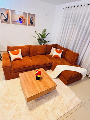 Una sala moderna con un sofá seccional marrón, una mesa de centro de madera con un florero y un ventanal con cortinas blancas.