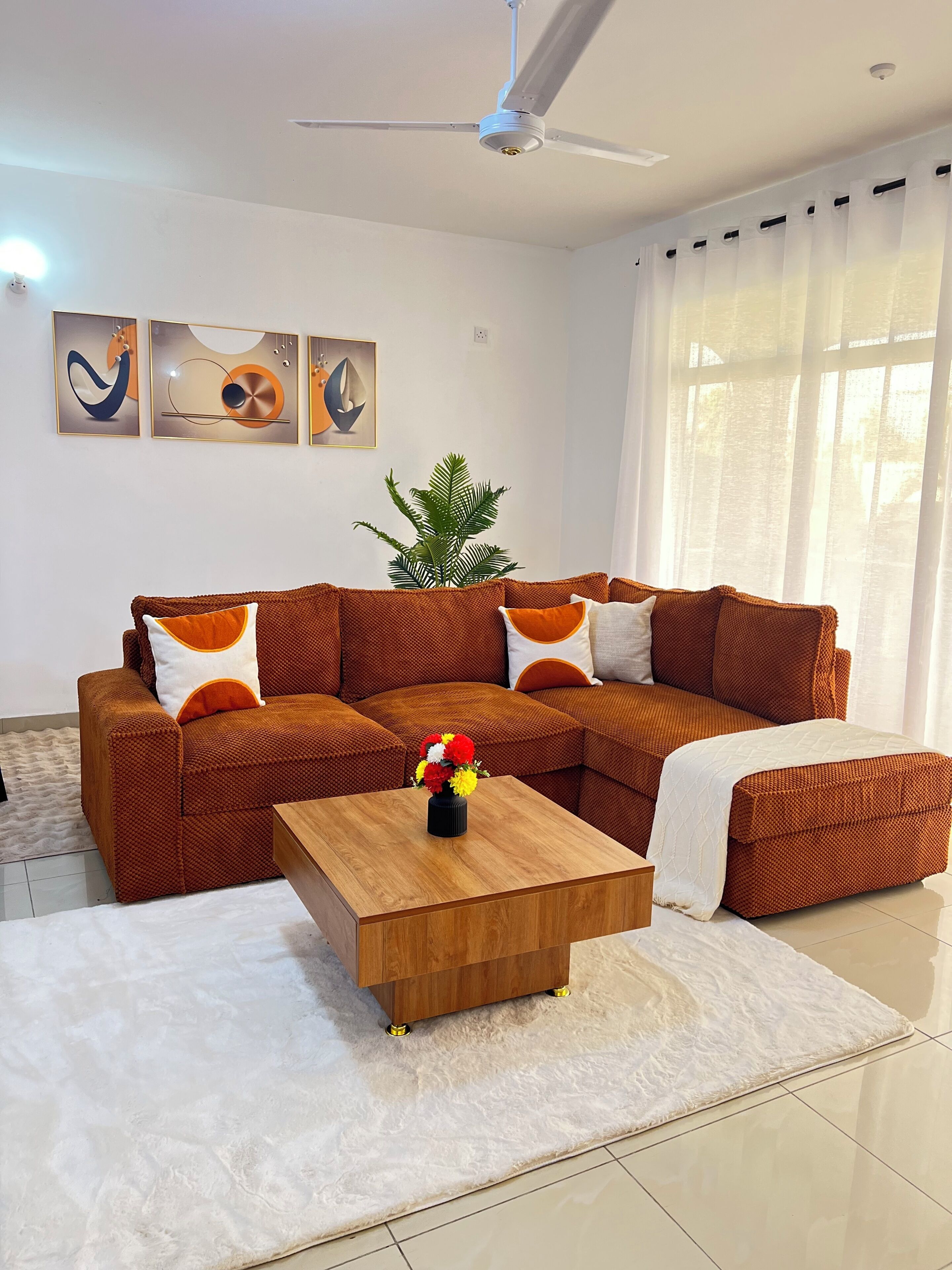Ein Wohnzimmer mit einem braunen Ecksofa, einem Couchtisch aus Holz und einem weißen Teppich. An der Wand hängen drei abstrakte Gemälde, ein Deckenventilator und eine Pflanze.