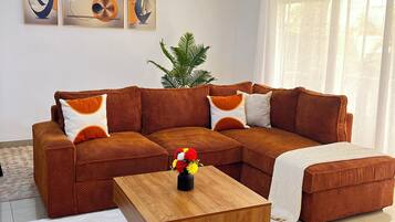 Ein Wohnzimmer mit einem braunen Ecksofa, einem Couchtisch aus Holz und einem weißen Teppich. An der Wand hängen drei abstrakte Gemälde, ein Deckenventilator und eine Pflanze.
