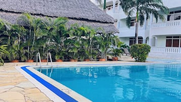 Une piscine bordée de pierres, une structure à toit de chaume et un immeuble à plusieurs étages doté de balcons.