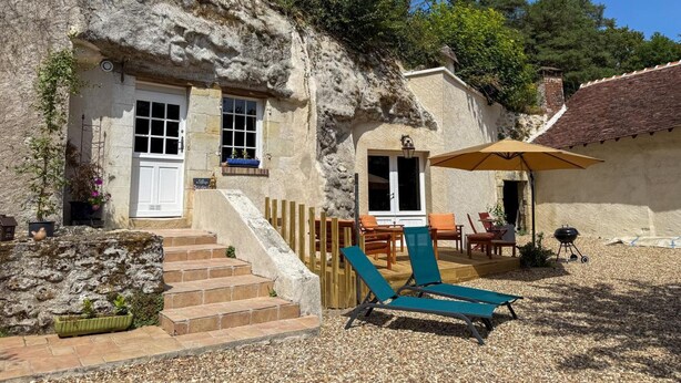 A rustic stone building with a wooden staircase leading to a door, outdoor seating area with a table and chairs, and a barbecue grill. - Unique troglodyte cave - 4 guests (Nazelles-Négron)