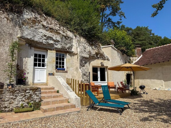 Ein rustikales Steinhaus mit einer Holztreppe, die zu einer Tür führt, einem Außenbereich mit Tisch und Stühlen sowie einem Grill.